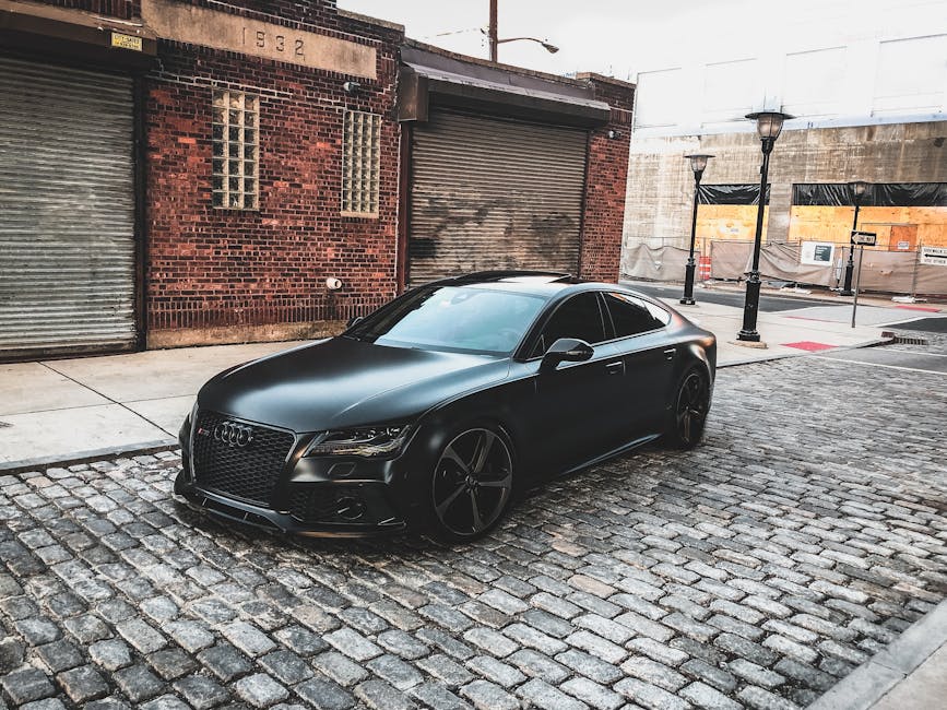 A sleek black car parked on a cobblestone street in an urban setting, featuring brick buildings.